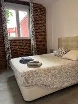 Dormitorio doble con pared de ladrillo visto, ventana con cortinas de hojas y cama con toallas dobladas